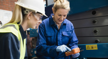 In a workshop. An employer dressed in overalls shows a high school learner in a hard hat a piece of equipment.