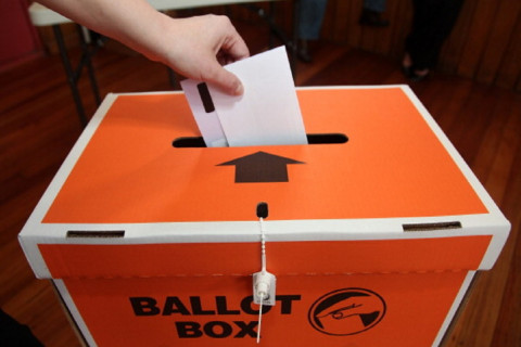 A hand places a voting ballot in an orange ballot box