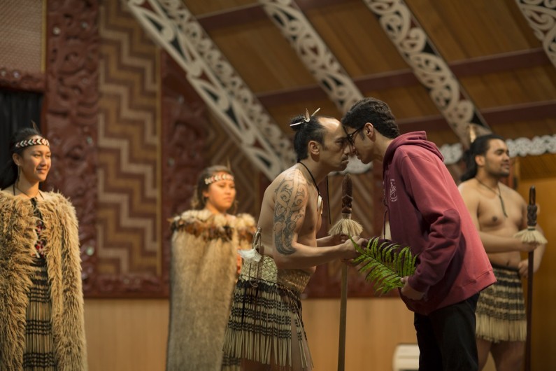 In the meeting house of a marae a man hongi with a young man.