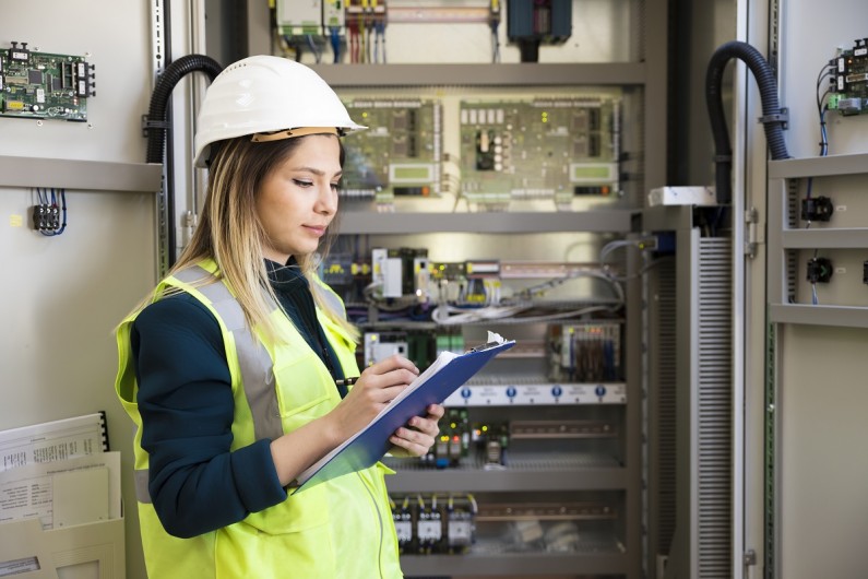 Engineer standing in front of a control panel writes notes on a clipboard