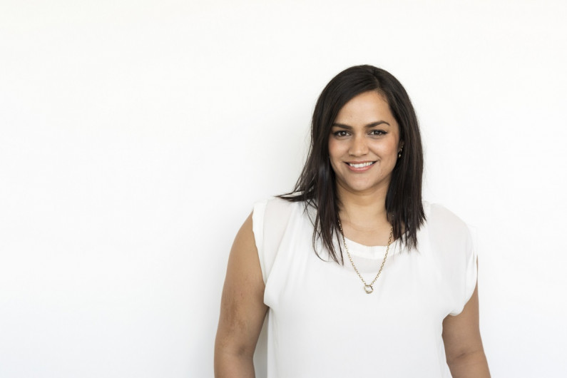 A woman stands in front of a white wall and smiles at the camera