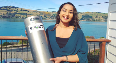 Jo Mohan stands in front of Wellington Harbour holding a milk churn and smiling