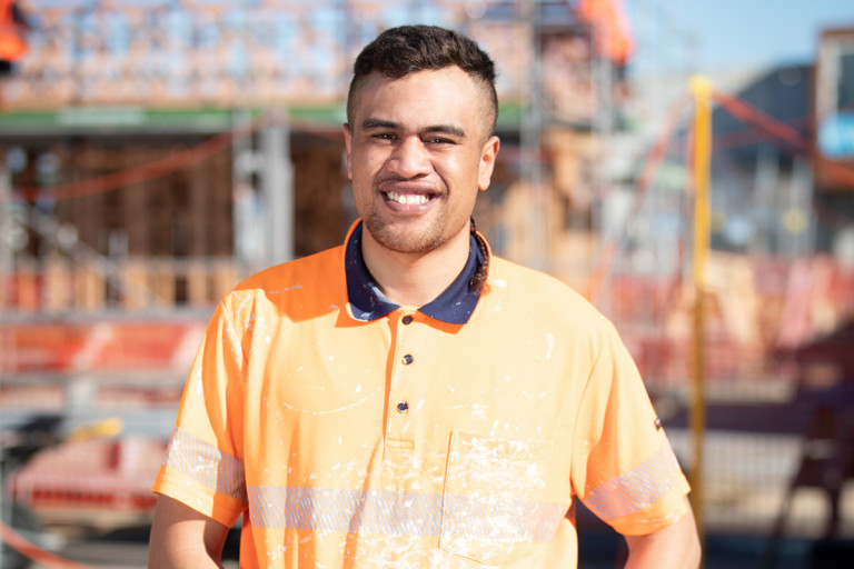 Young man on building site is wearing safety gear and smiling at the camera.