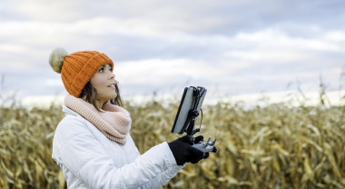 A woman holds the controls for a drone she is flying over corn fields.