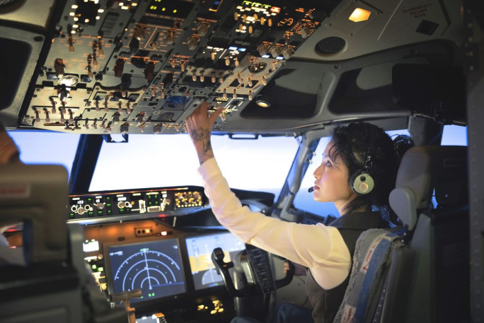 A  female pilot sits in an aircraft cockpit flipping a switch on the control panel on the cockpit ceiling 