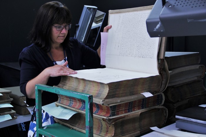 Susan Jenkins, archivist, looks at a very large historical volume on a stack of similar books on a trolley near a scanner