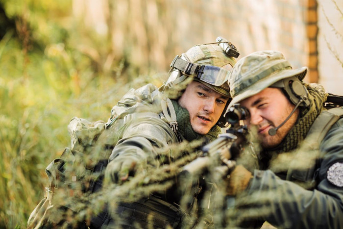 A male solider and officer both wearing camouflage and lying  in long grass have their heads pressed together as they both look down the barrel of a rifle. The officer is pointing at a target. 