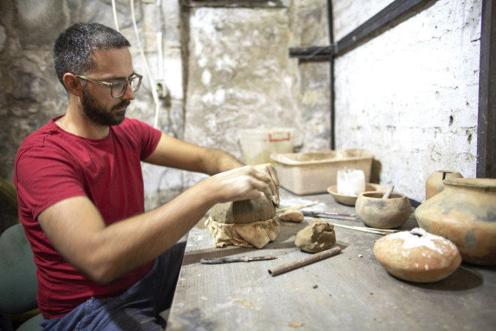 A man sits at a workbench making an object out of clay