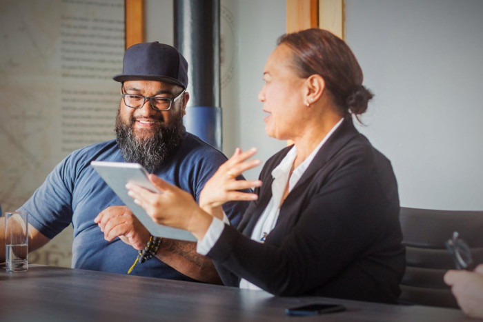 A man and woman sit at a table. She's wearing a formal jacket and white shirt, he's dressed causally. She's showing him what's on a tablet in her hand 