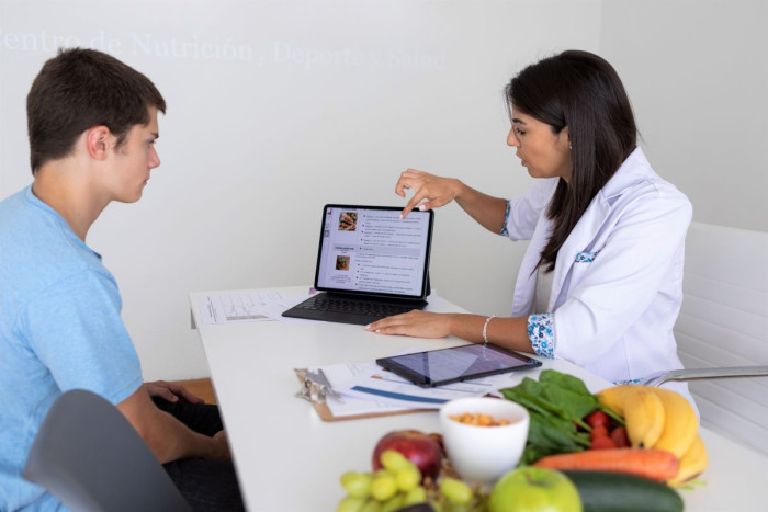 A female dietitian sits across a table from a teenage boy. Vegetables are piled on the table. She's pointing to a diagram about food groups on a tablet