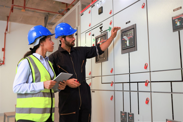 Two electrical engineers, a man and a woman, stand at an electricity control board. The man is reaching toward a switch on the board and the woman is holding a tablet. They both wear high viz vests and safety helmets