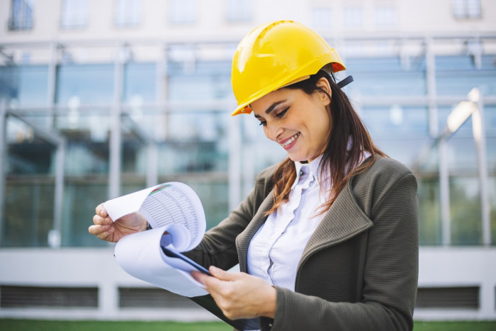 A female facilities manager wears a yellow hard hat and looks at her clipboard