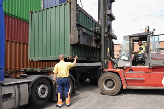 A man signals to a forklift operator who is manoeuvring a forklift to place a shipping container onto the back of a truck