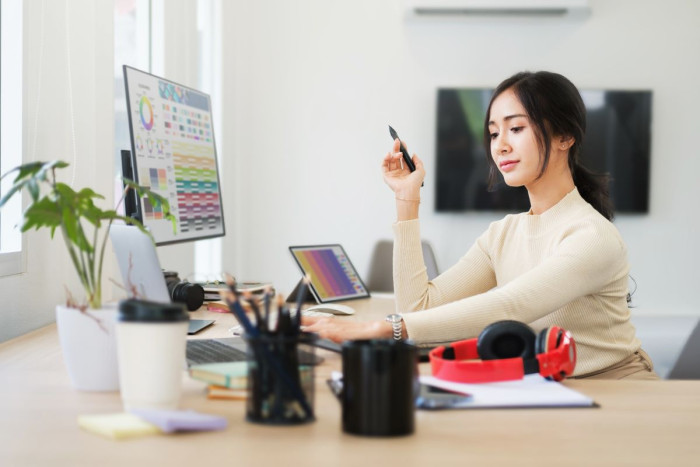 A woman sits at an office desk looking at a computer with a colourful display. On her desk is a tablet also showing rainbow colours 