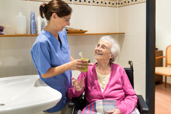A health care assistant in a uniform hands a glass and toothbrush to an elderly woman who is in a wheelchair with a rug over her knees, in a bathroom 