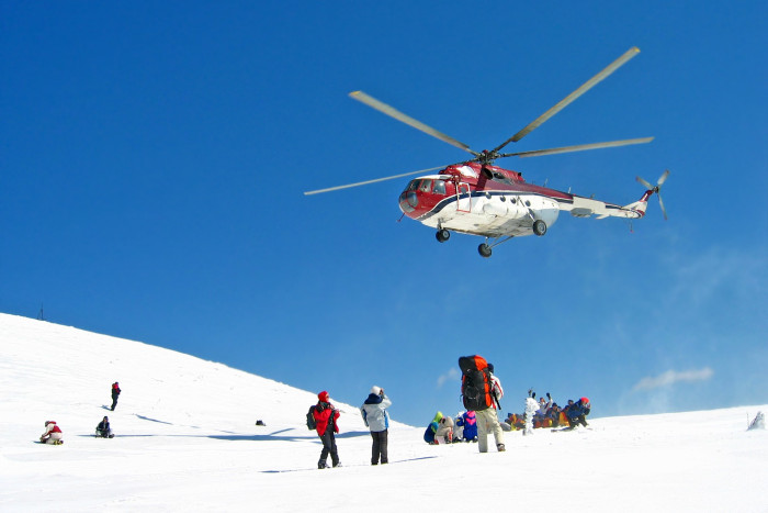 A helicopter in the air above a snowy hillside where people in outdoor clothing are waiting