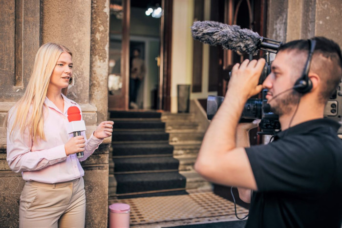 A woman speaks into a microphone she's holding while a camera person films her outside a building