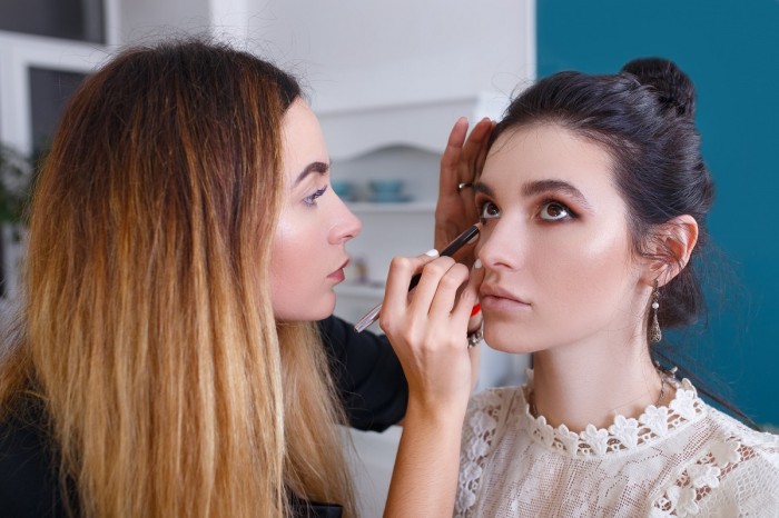 A make-up artist holds a model's head with one hand and applies eyeliner with the other