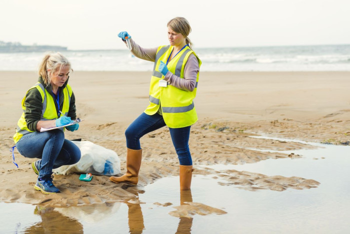 Two women are at the beach wearing high-vis vests. One is holding up a test tube and the other is kneeling down writing notes 