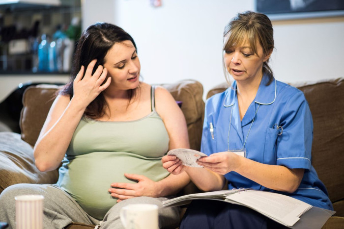 A pregnant person and a midwife sit on a soft in a home. The midwife is holding a circular tool which calculates due dates for babies 
