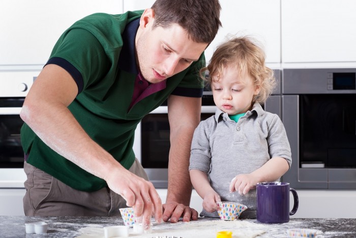 A young man in a home kitchen is showing a toddler boy how to bake cupcakes