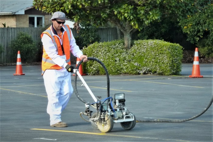 A man wearing a high-viz jacket and earmuffs paints a yellow line with a painting machine in a car park 