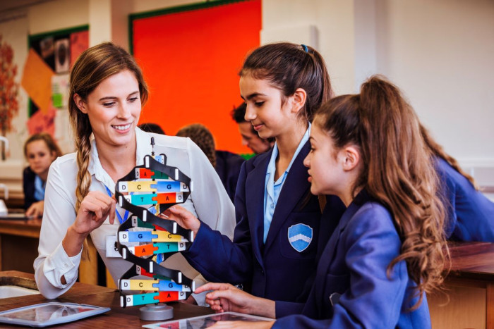 A female science teacher and two teenage students in school uniform look at a model of a double helix