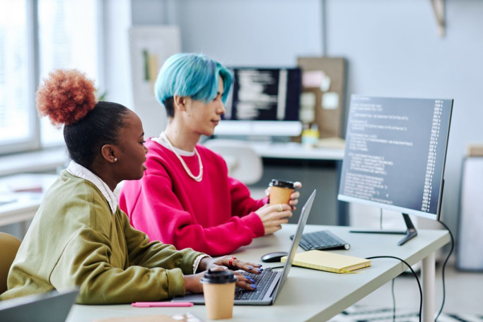 Two software developers, one with blue hair and a pink sweatshirt and the other with pink hair and a green sweatshirt, both with coffee, sit at a desk looking at code on a computer screen