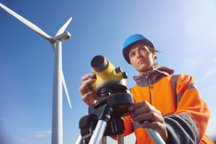 A woman in a hard hat and a fluro jacket stands in front of a builder's level with a wind turbine in the background 