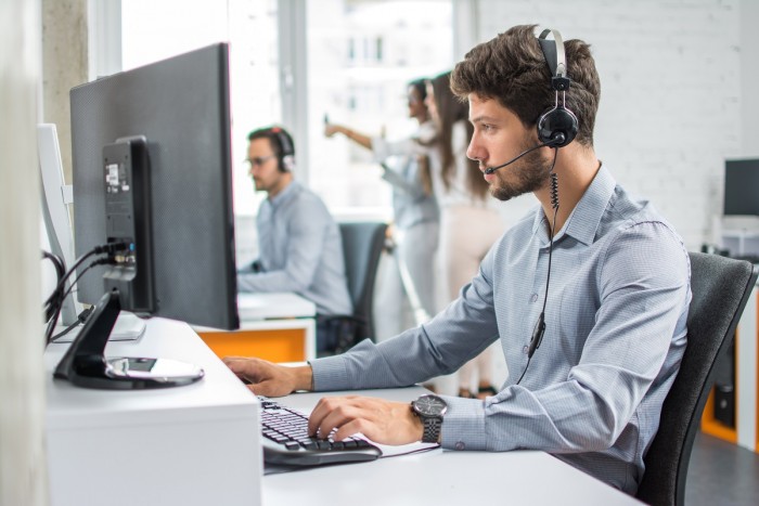 A young man wearing a headset checks the computer as he talks to a customer on the phone