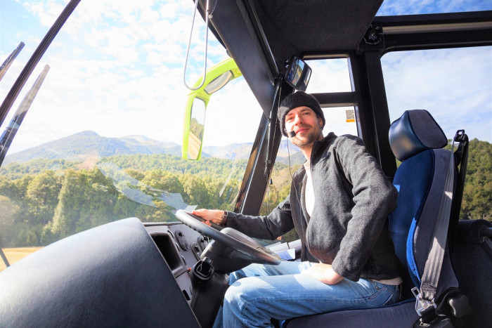 A tour guide who is also a bus driver sits in the driver's seat of a bus wearing a microphone and turning towards his passengers