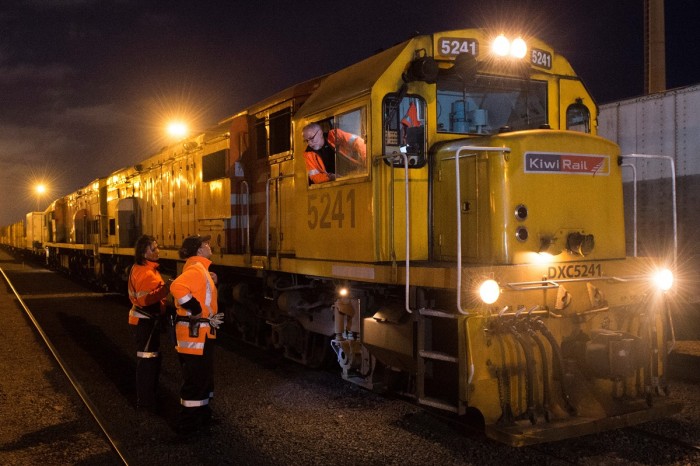 A train driver in a railway yard at night leans out of his cab to talk to two workmates standing below 