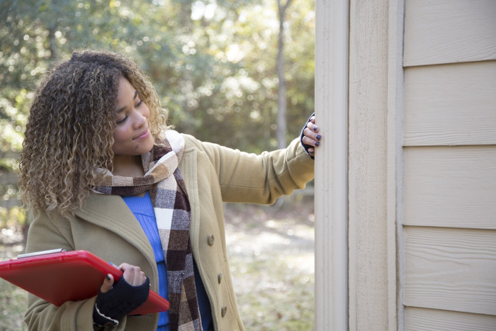 A property valuer has her hand on the side of the outside of a house she's inspecting. She's holding a clipboard.