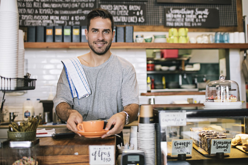 A man smiles facing the camera while he offers a cup of coffee. He is behind the counter in a cafe surrounded by cakes and a coffee machine.
